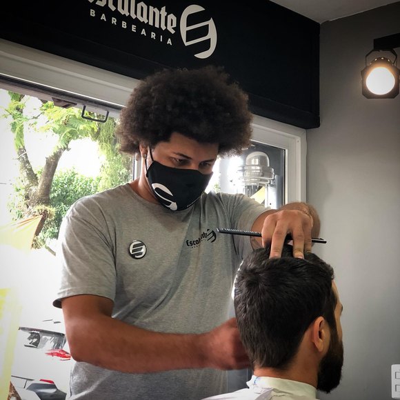 Barbeiro trabalhando cabelo crespo com tesoura, na cadeira junto à janela.