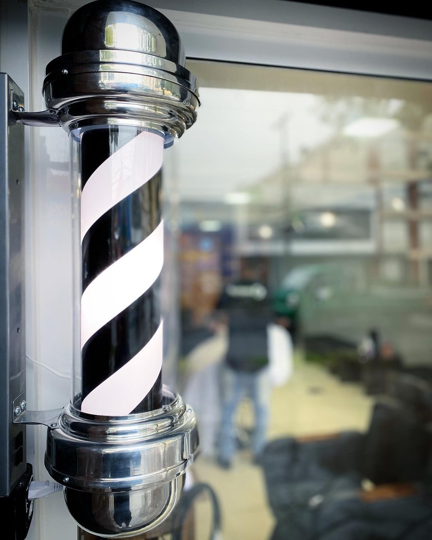 O poste de barbeiro cromado na porta da Escalante, com a barbearia refletida no vidro atrás.