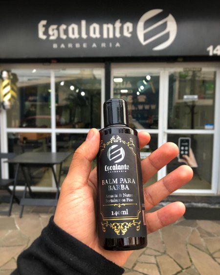 Frasco do balm para barba da Escalante na mão, em frente à fachada.