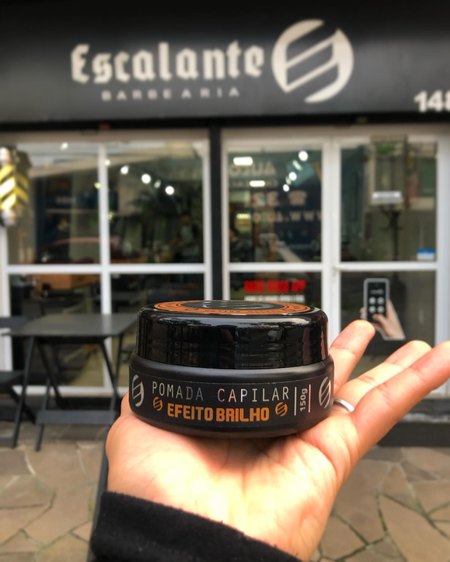 Pote de pomada capilar efeito brilho da Escalante na palma da mão, em frente à fachada.