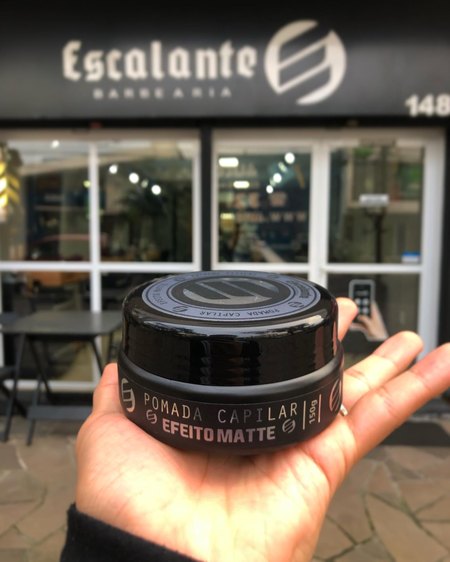 Pote de pomada capilar efeito matte da Escalante na palma da mão, em frente à fachada.