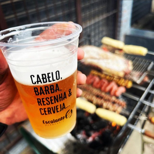 Copo da barbearia com cerveja, escrito “cabelo, barba, resenha & cerveja”, na frente da churrasqueira.